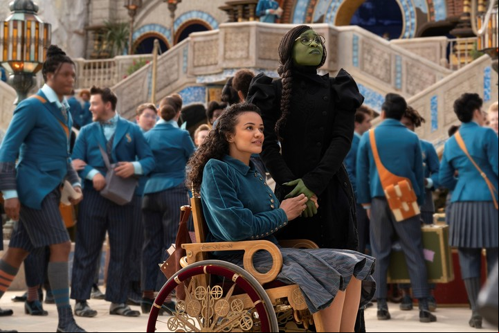 Marissa Bode, sentada, como la hermana de Elphaba, llegando a la Universidad de Shiz, junto a Cynthia Erivo.
